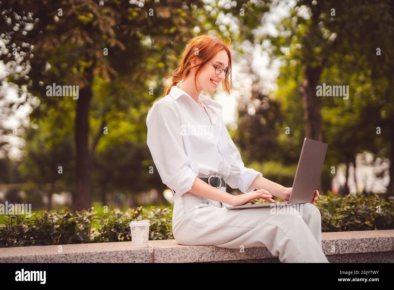Beautiful redhead young woman sitting in the park and using laptop ...