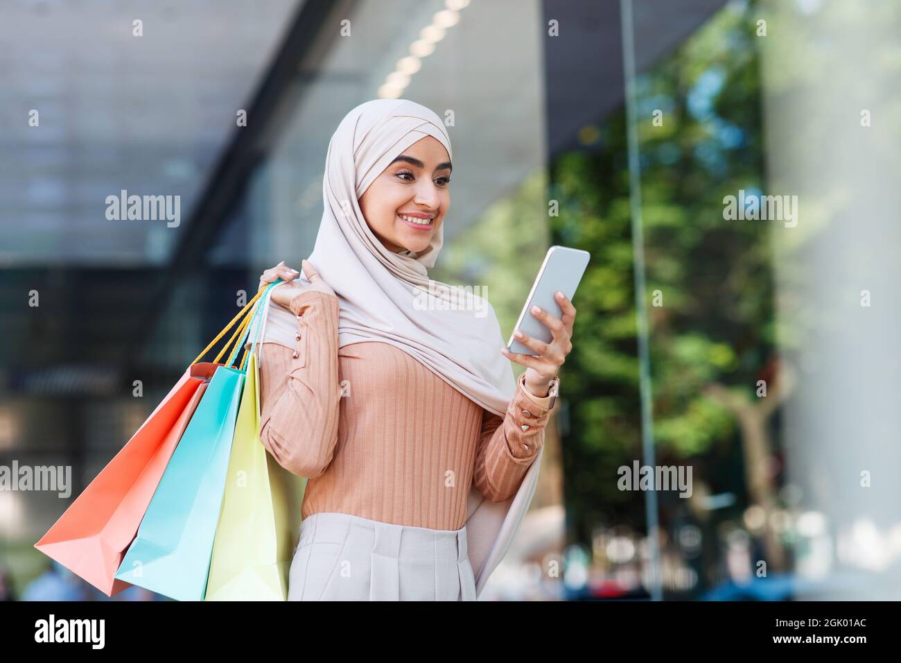Satisfied cute young middle eastern female in hijab with many packets, texting message on smartphone near mall Stock Photo