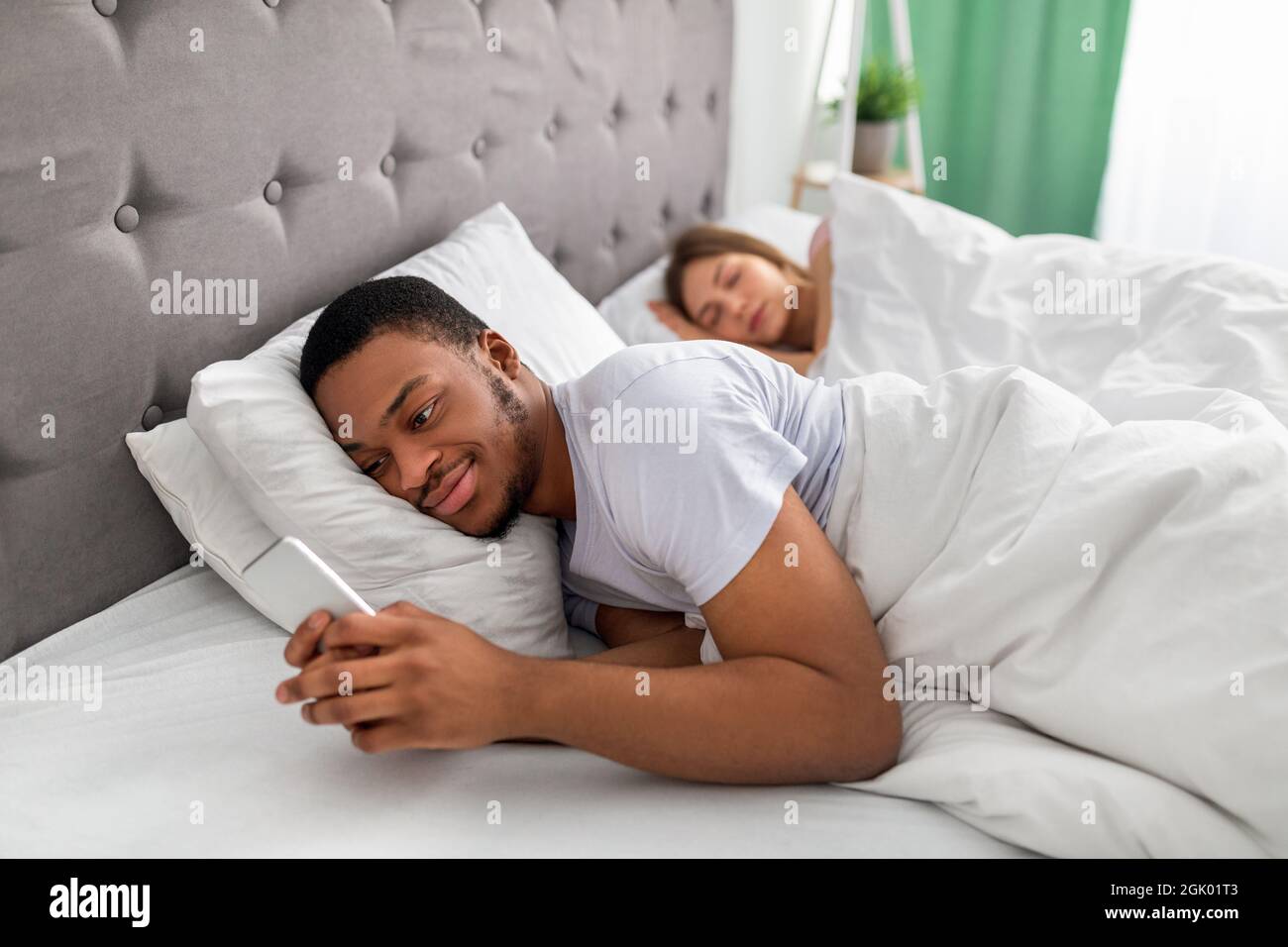 Young black guy lying on bed and using mobile phone to text his lover while his wife is asleep. Relationship infidelity Stock Photo