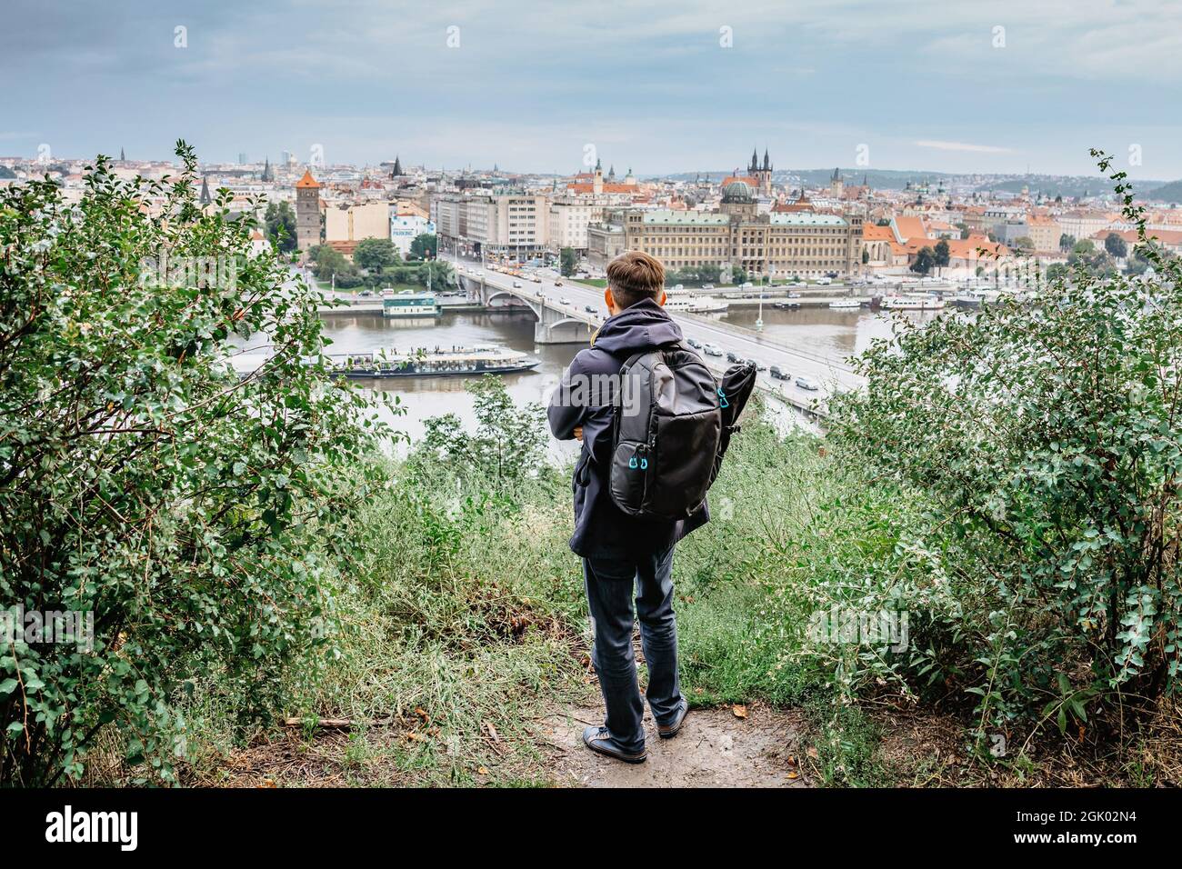Young male backpacker enjoying view of Prague city skyline and Vltava river from Letna Park,Czech Republic.Prague panorama on cloudy rainy day.Travel Stock Photo