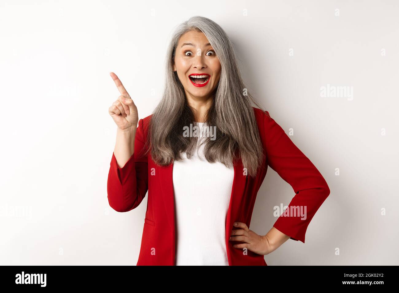 Business concept. Cheerful asian businesswoman with grey hair, wearing red blazer and makeup, pointing upper left corner and smiling amazed, white Stock Photo