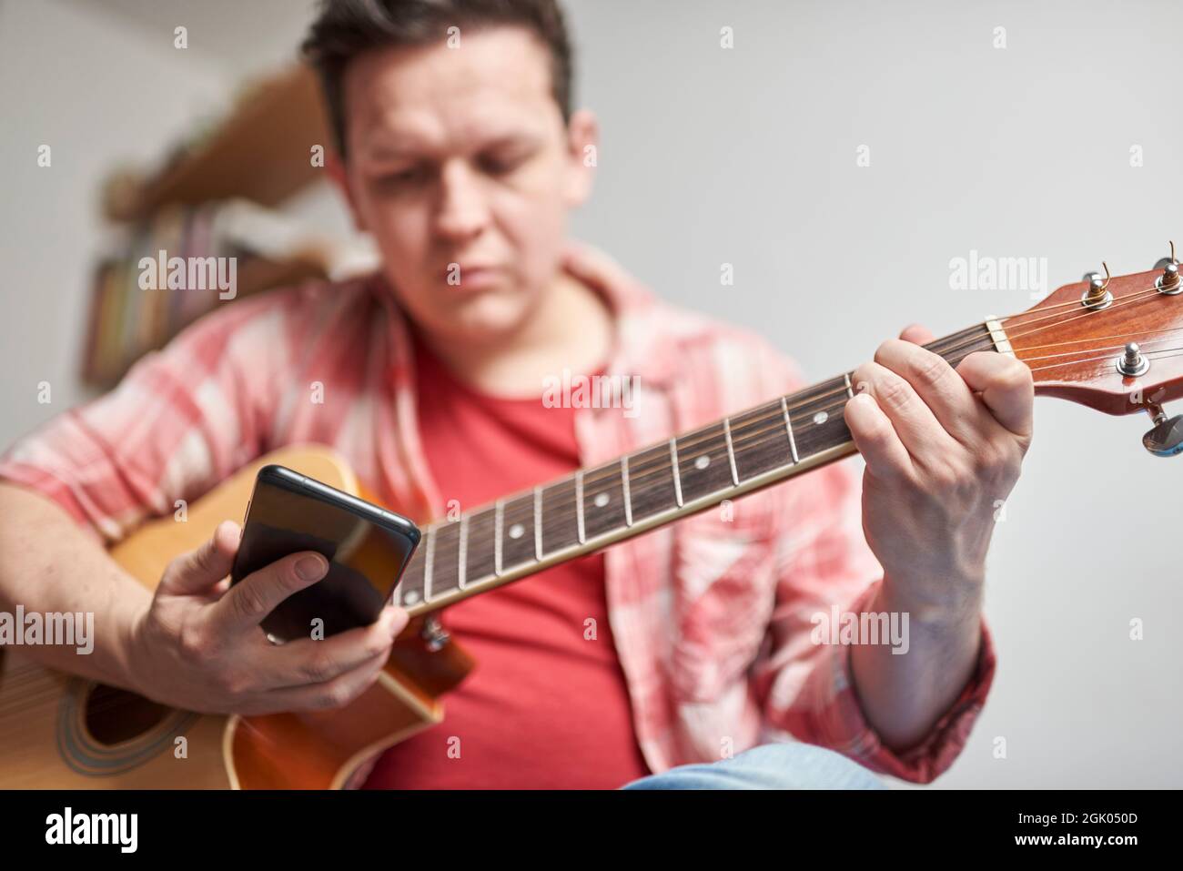 Young man learning to play guitar at home with the help of his smartphone. Concepts of virtual education, hobbies and music. Focus in hands. Stock Photo