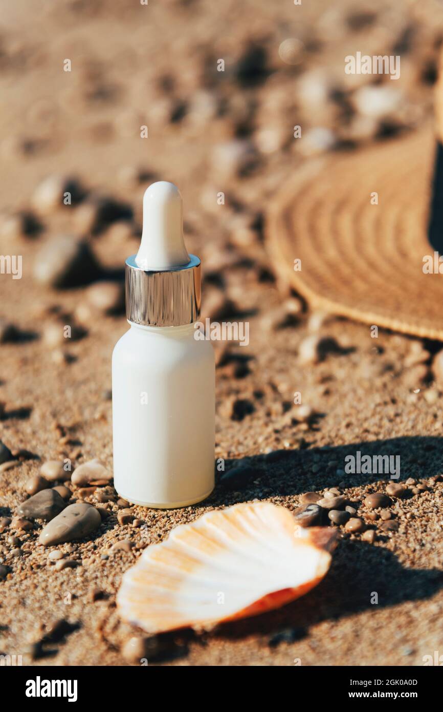Cosmetic moisturizer, oil or serum bottle in beach sand next to seashell and straw hat. Natural