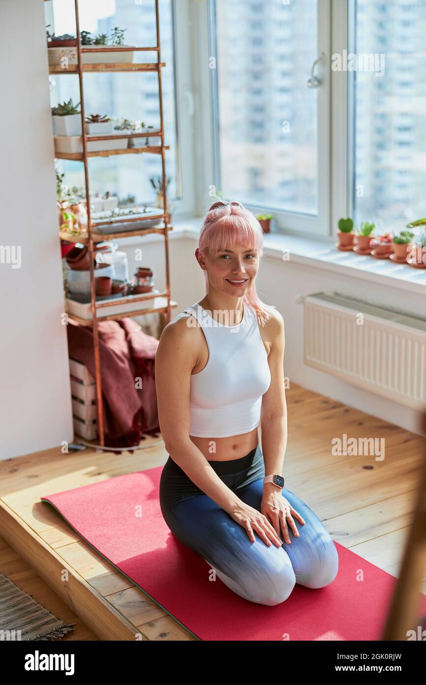 Pretty lady with pink hair in tracksuit sits on knees before sports training at home Stock Photo