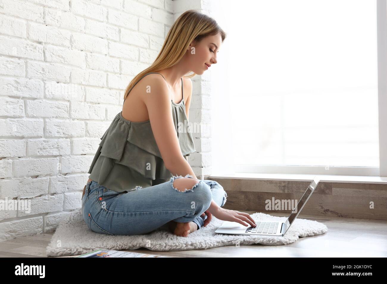 Young girl sitting on rug and using laptop near big window in room