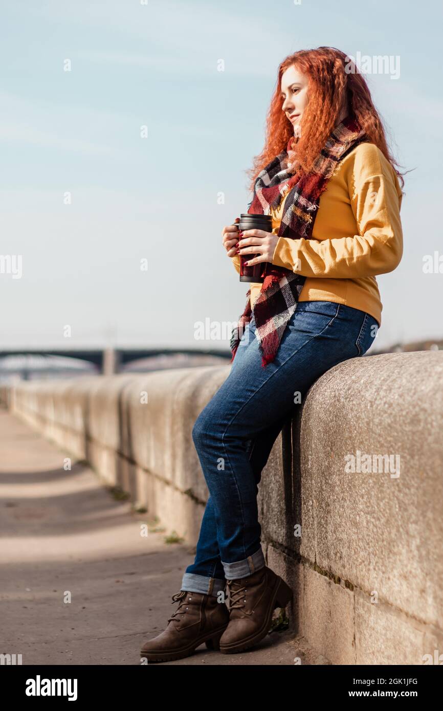 Young woman in yellow sweater sits on embankment and drinks coffee from reusable mug. She is enjoying autumn sunny day. Eco-friendly lifestyle, relaxa Stock Photo