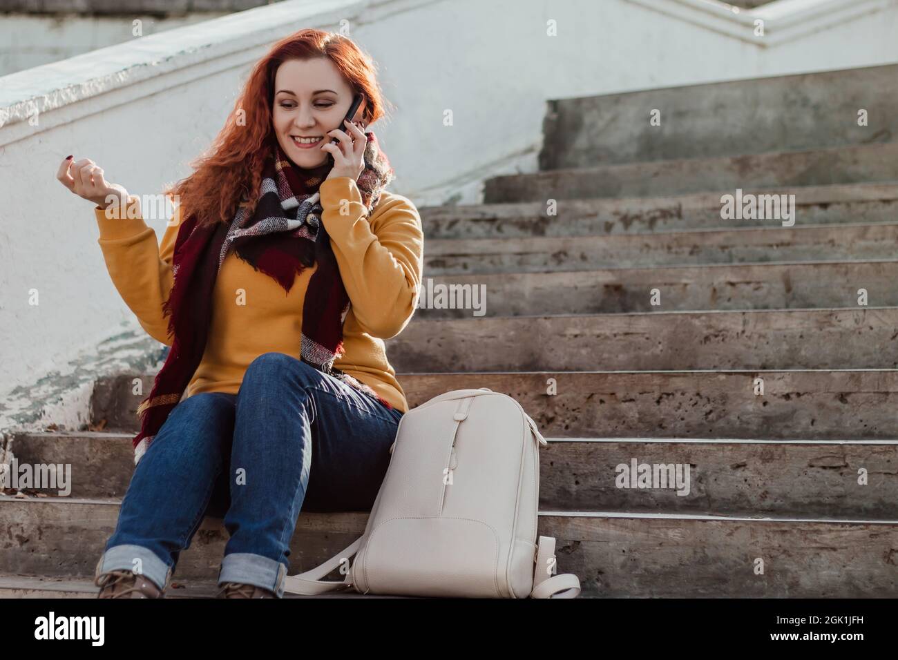 Young red-haired woman sitting on stairsand talking on phone. Lady in yellow sweater uses smartphone on sunny day. Lifestyle, copy space. Stock Photo