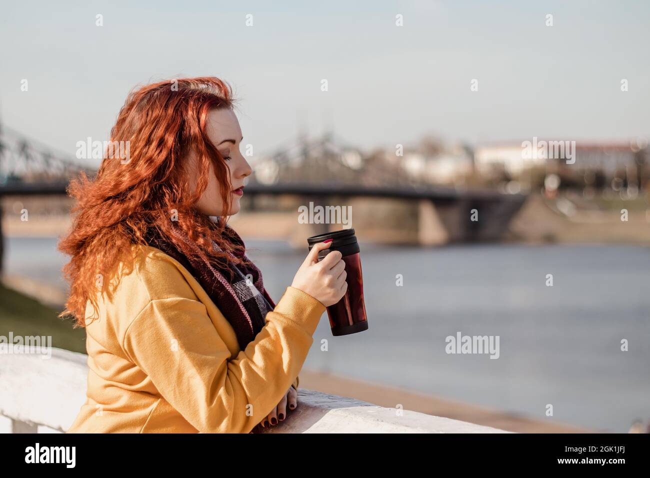 Young red-haired woman in yellow sweater holds thermo mug with coffee in her hands. Enjoy the river view on sunny autumn day. Eco-friendly lifestyle, Stock Photo