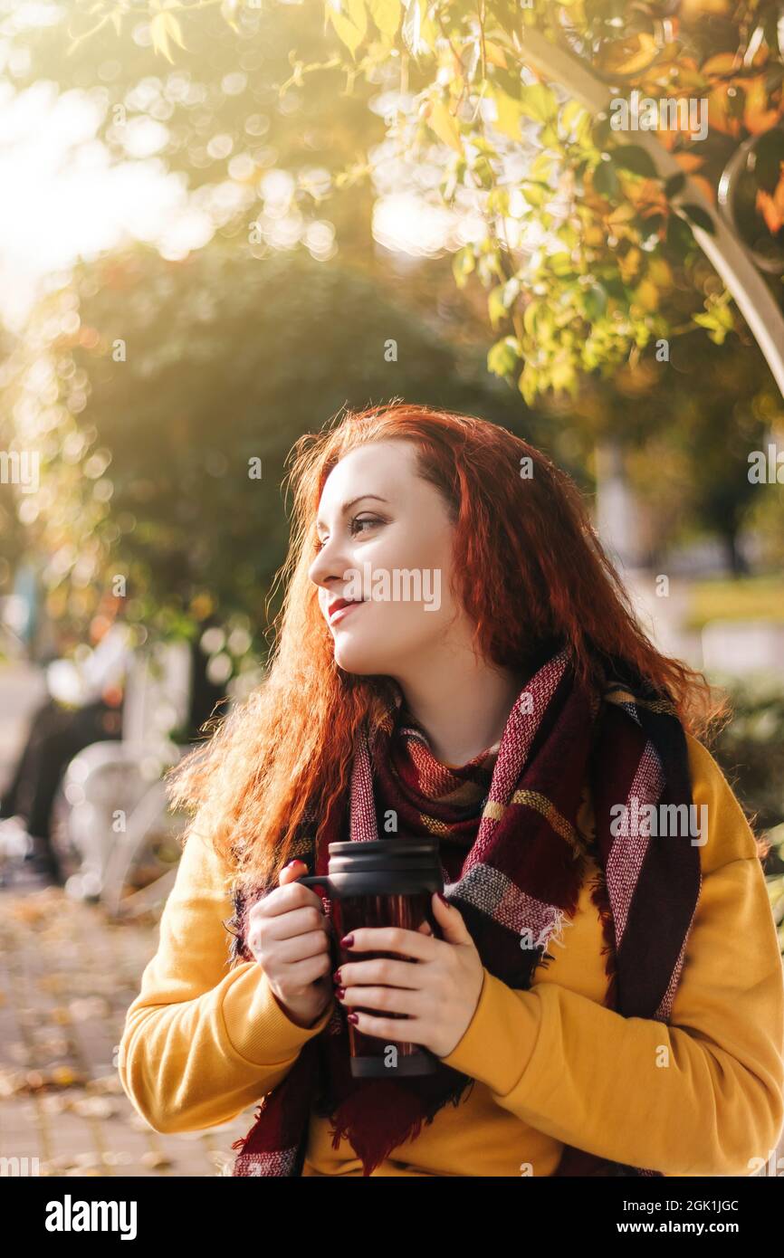 Young red-haired woman holds reusable mug in her hands. She sits on park bench and is wearing yellow sweater. Lifestyle on a sunny autumn day. Stock Photo