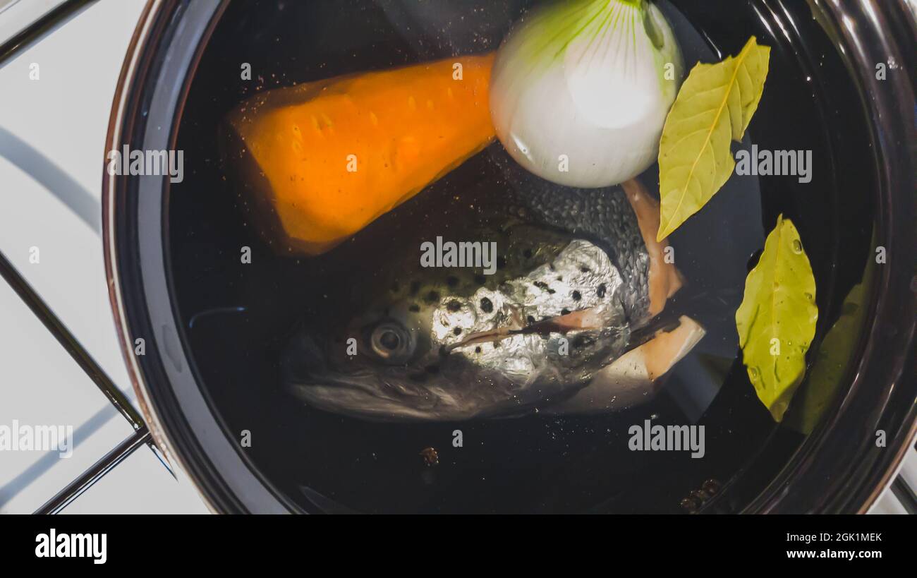The broth from the head of salmon, onions, carrots is cooked in a saucepan on a white gas stove. Together with bay leaves and allspice peas. Top view, close-up. Horizontal photography. Stock Photo