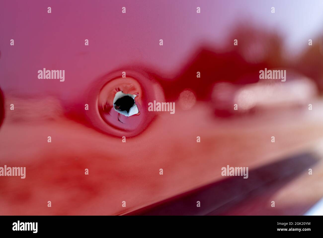 Close up shot of a bullet hole on within the passenger side door of a red car with a reflection of a car visible. Stock Photo