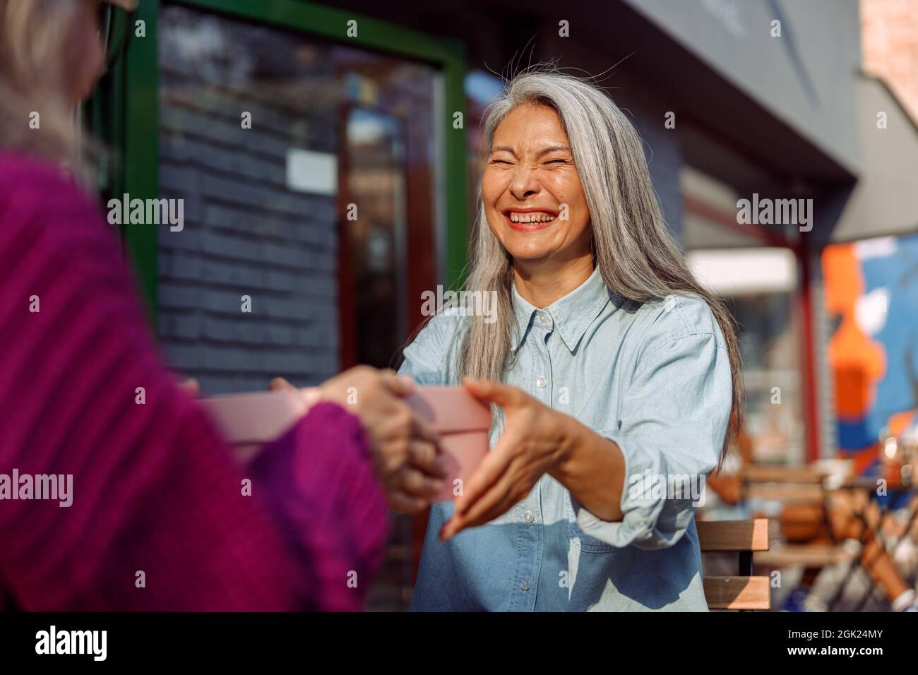 Cheerful mature Asian woman with friend hold gift box sitting at small table in cafe Stock Photo