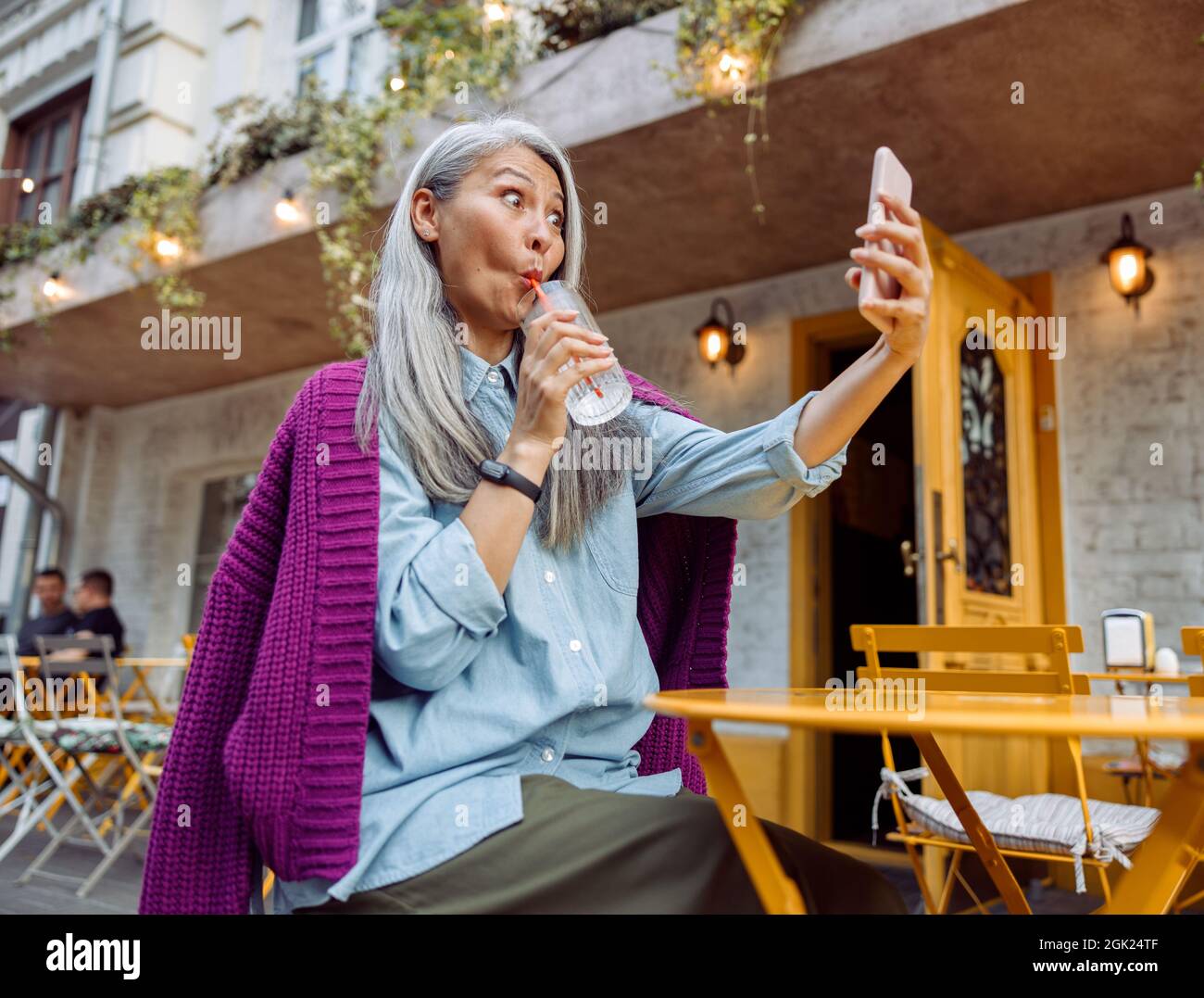 Funny senior Asian woman drinks water through straw taking selfie at cafe table outdoors low angle view Stock Photo