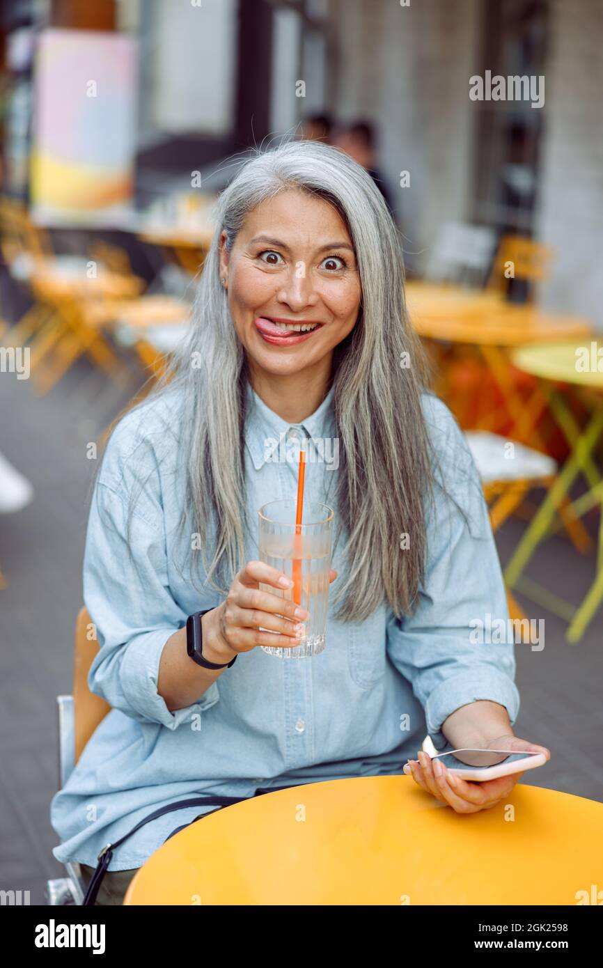 Mature Asian woman makes funny face sitting at small table on outdoors cafe terrace Stock Photo