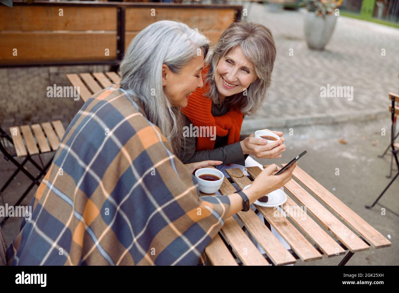 Mature Asian woman shows mobile phone to best friend in street cafe on autumn day Stock Photo