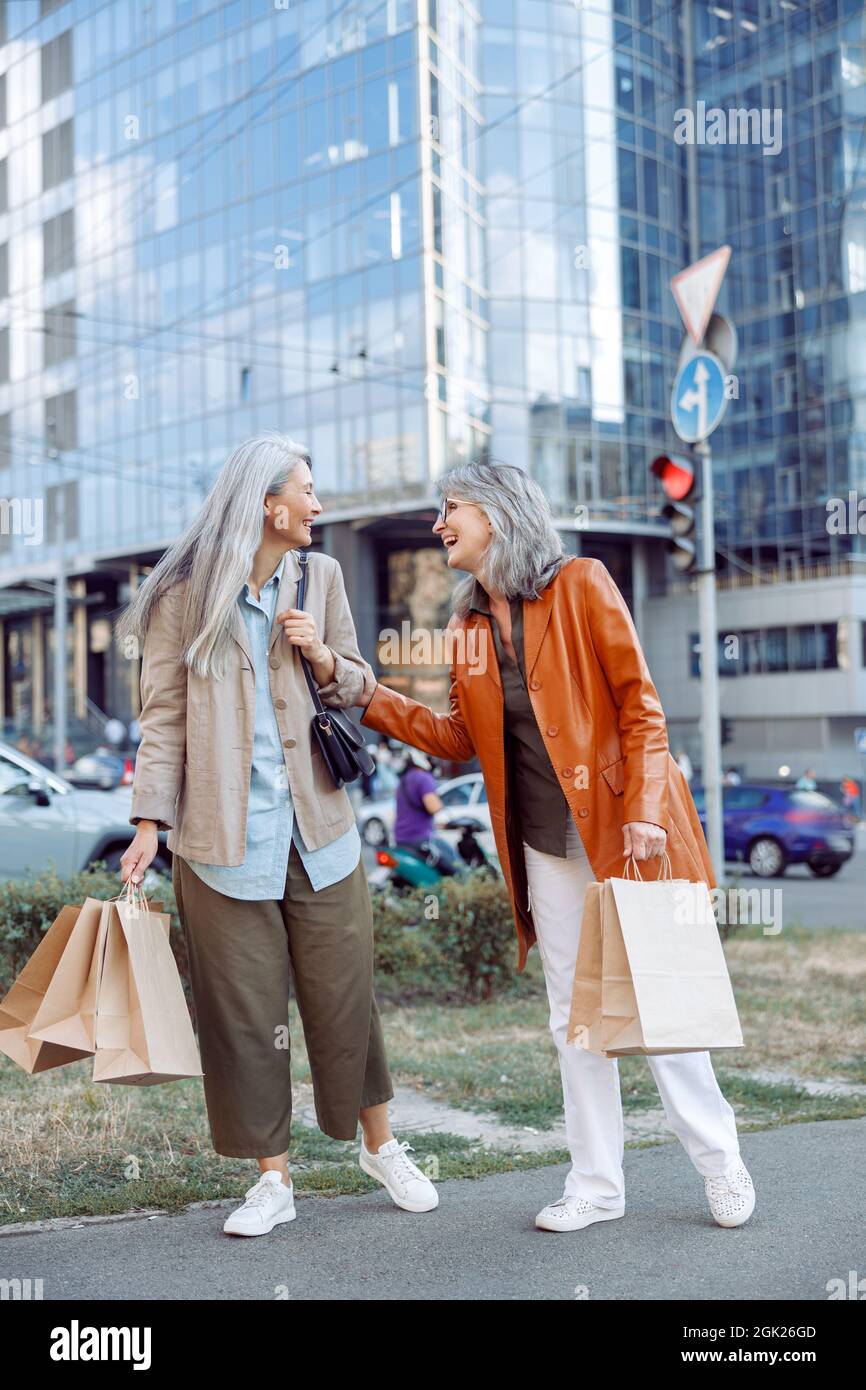 Mature woman with shopping bags touches Asian lady elbow laughing on ...