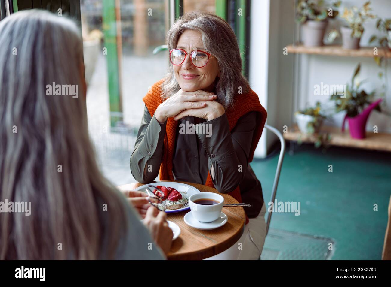 Grey haired woman with glasses looks at friend sitting at small table in cozy cafe Stock Photo