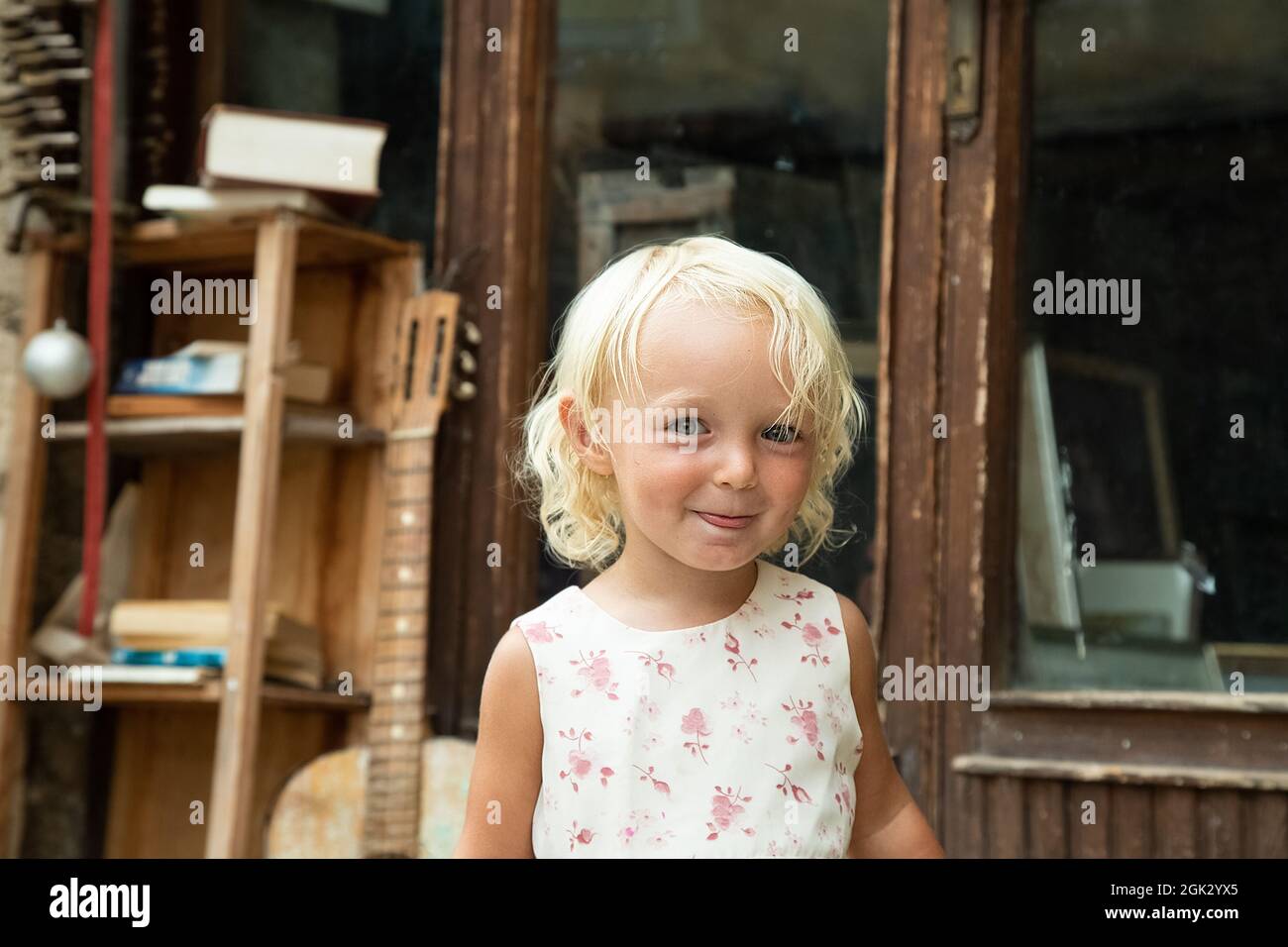 Little blond blue eye girl in white dress with pink flowers portrait. Tricky funny child. Retro style picture. Vintage door with bookshelf and guitar Stock Photo