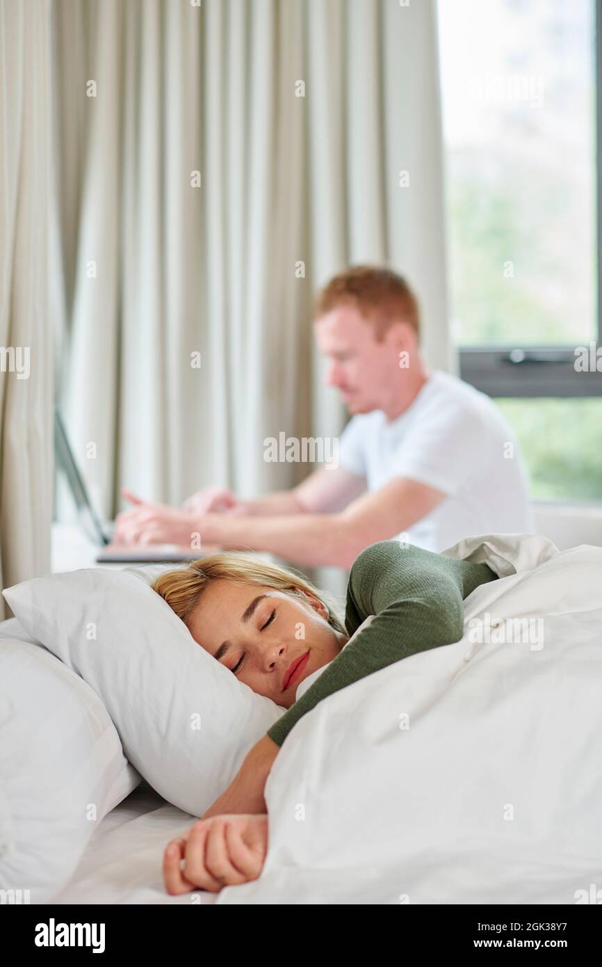 Beautiful young woman enjoying sleeping in comfy bed when her boyfriend is working on laptop in background Stock Photo