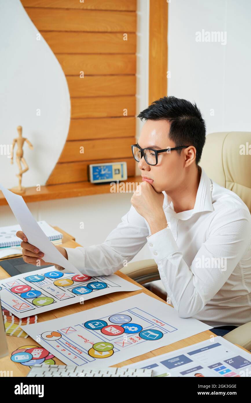 Contemplating young UX designer looking at dociment with mobile application layout he created Stock Photo