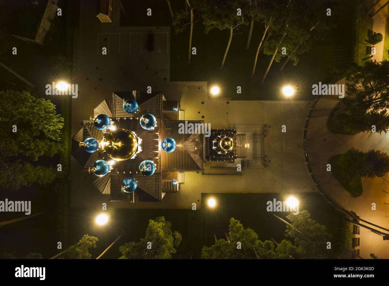Aerial view of illuminated church between pine trees at night. New public empty white chapel with gold and blue domes outdoors from above. Beautiful Religious, sacred place for ministries Stock Photo