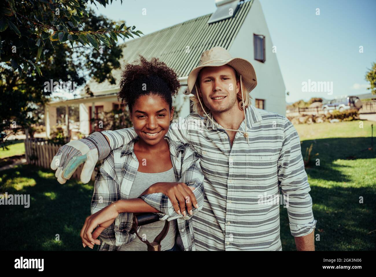 Mixed race young couple standing in luscious green fields in front of white picket fence home Stock Photo