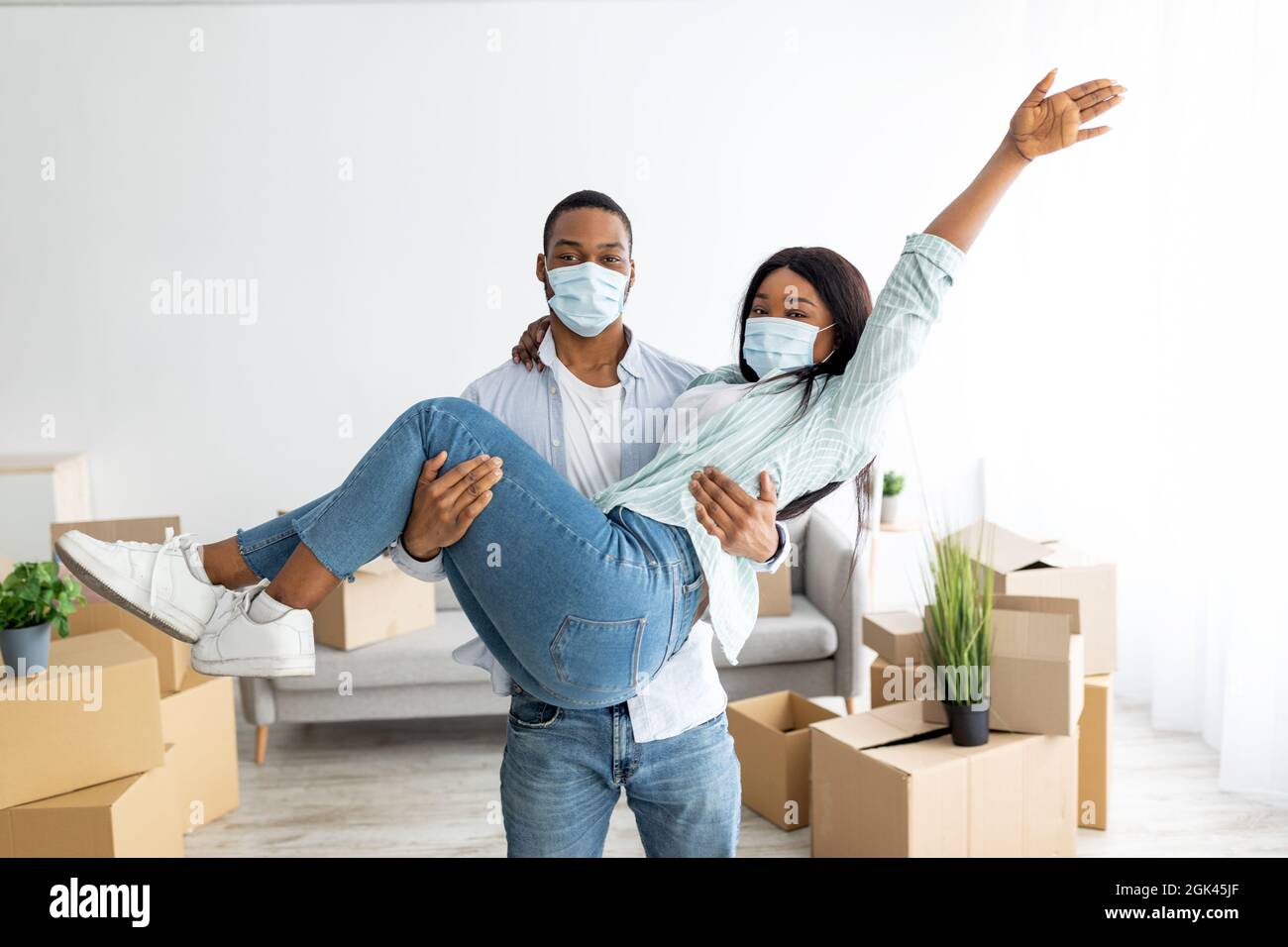 Celebrating moving day. Happy african american couple in protective masks having fun, man holding woman on hands Stock Photo