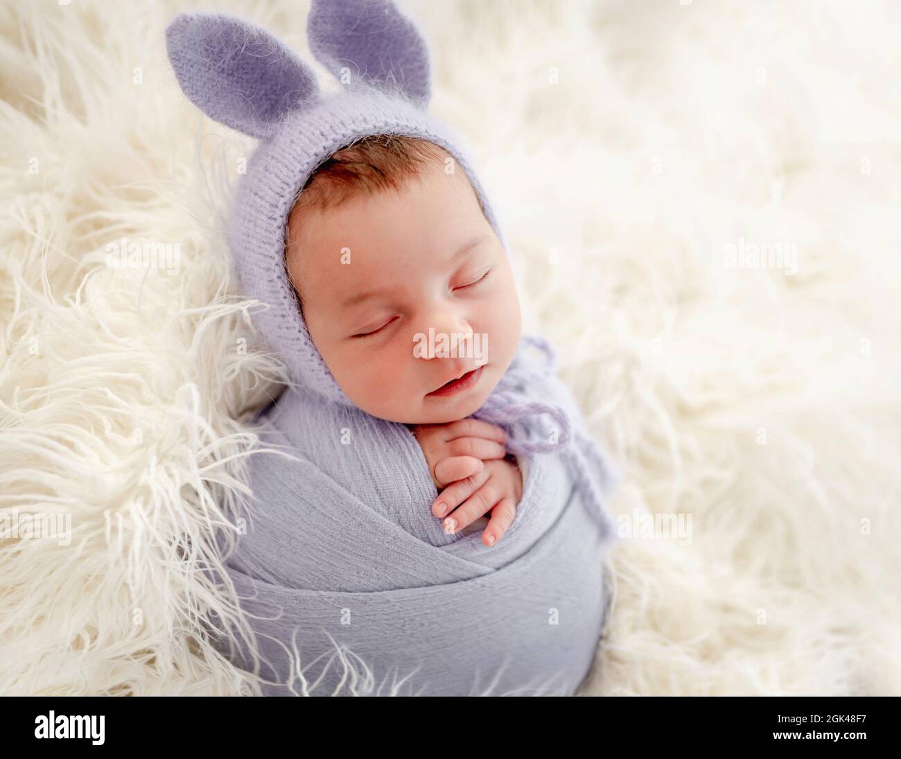 Little beautiful newborn baby girl swaadled in fabric and wearing hat with bunny ears sleeping on fur during studio photoshoot. Cute infant child kid Stock Photo
