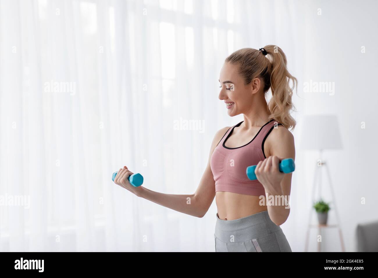 Cheerful european cute young blonde woman lifts up dumbbells at home, work out alone, takes care of body Stock Photo
