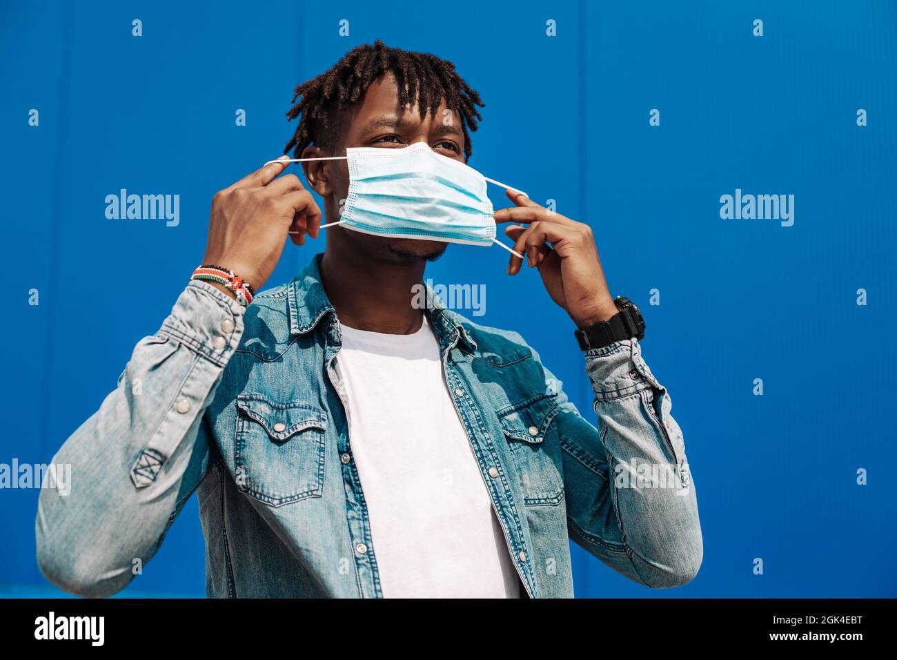 portrait of an afro american black man wearing a protective mask against coronavirus, on a blue background, guy puts a mask on his face Stock Photo