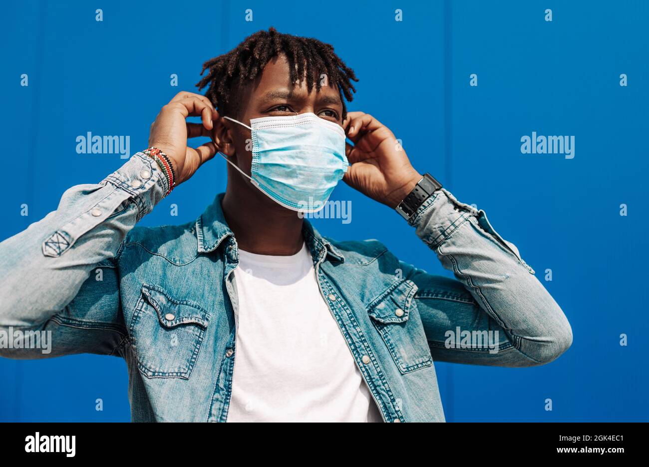 portrait of an afro american black man wearing a protective mask against coronavirus, on a blue background, guy puts a mask on his face Stock Photo