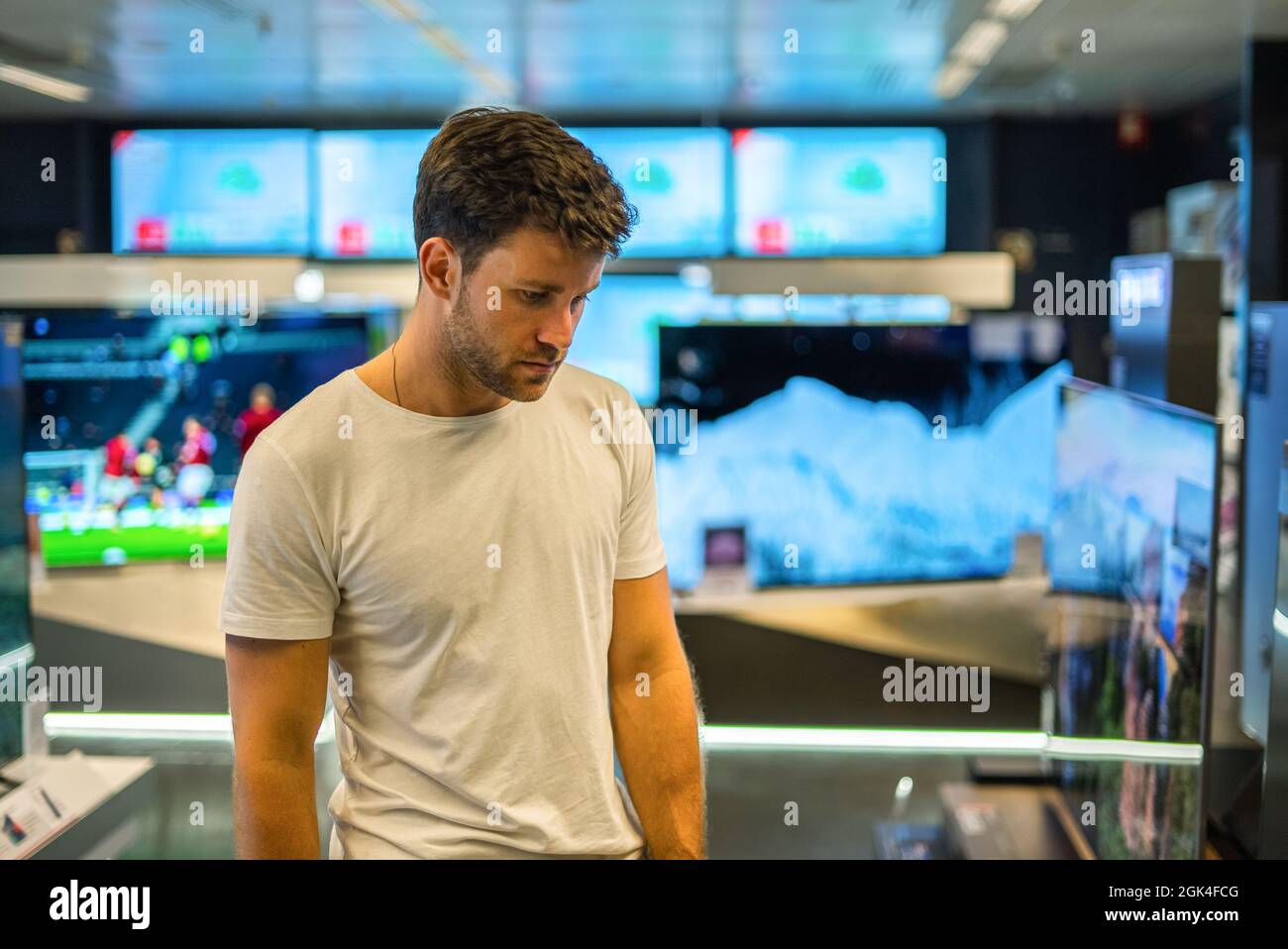 Serious attentive young bearded male customer in casual clothes standing in department with modern TV sets and examining goods while choosing purchase Stock Photo