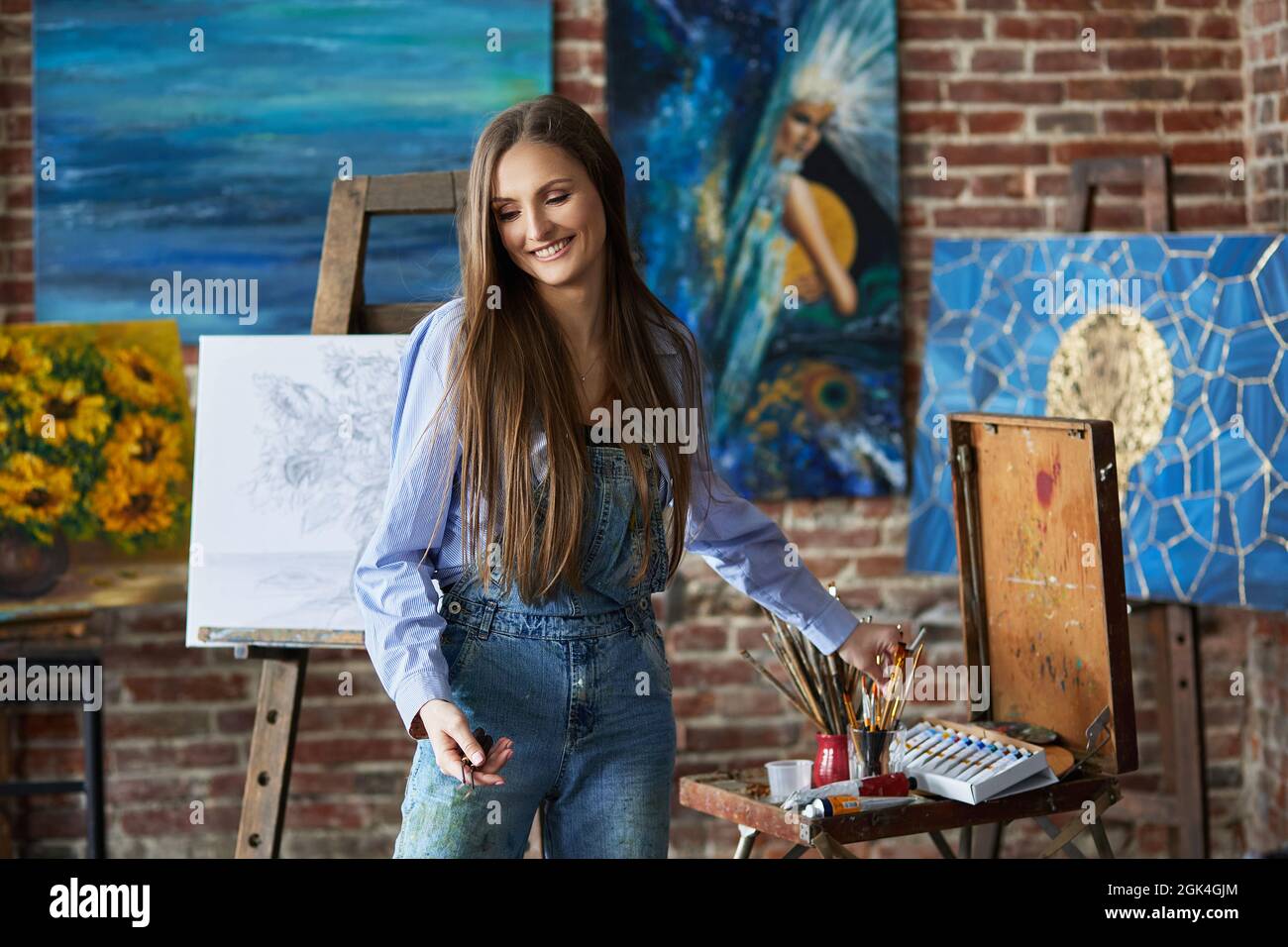 Portrait of young smiling female artist with her artworks in art studio ...