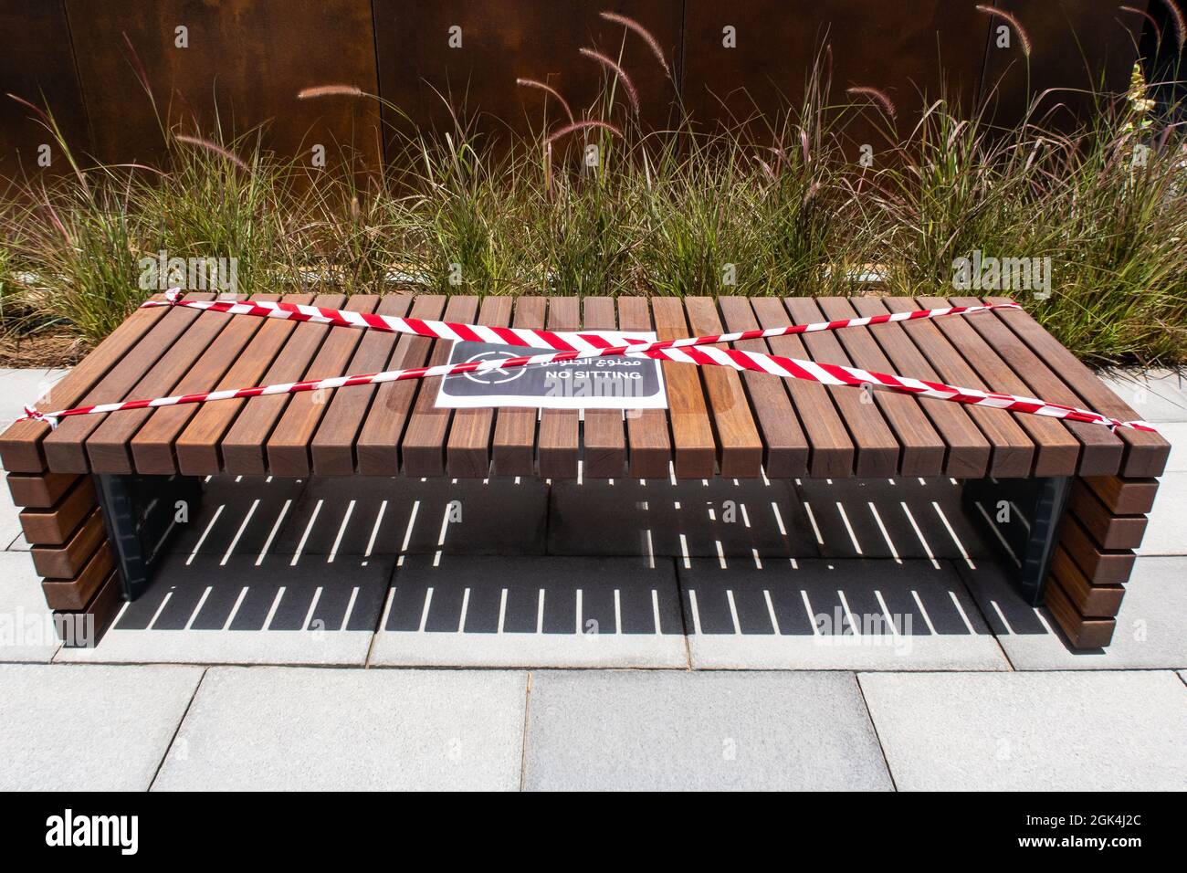 Wooden brown bench with 'No Sitting' sign in english and arabic and red and white barricade tape, safety measures due to coronavirus COVID-19 in Dubai Stock Photo