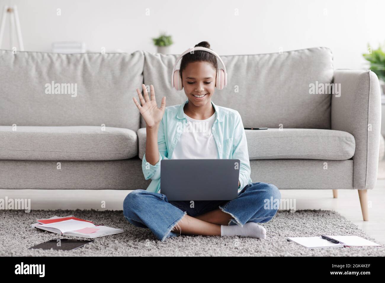 Cheerful teen black girl learns remotely, calls online and waves hand to laptop, sitting on floor at home Stock Photo