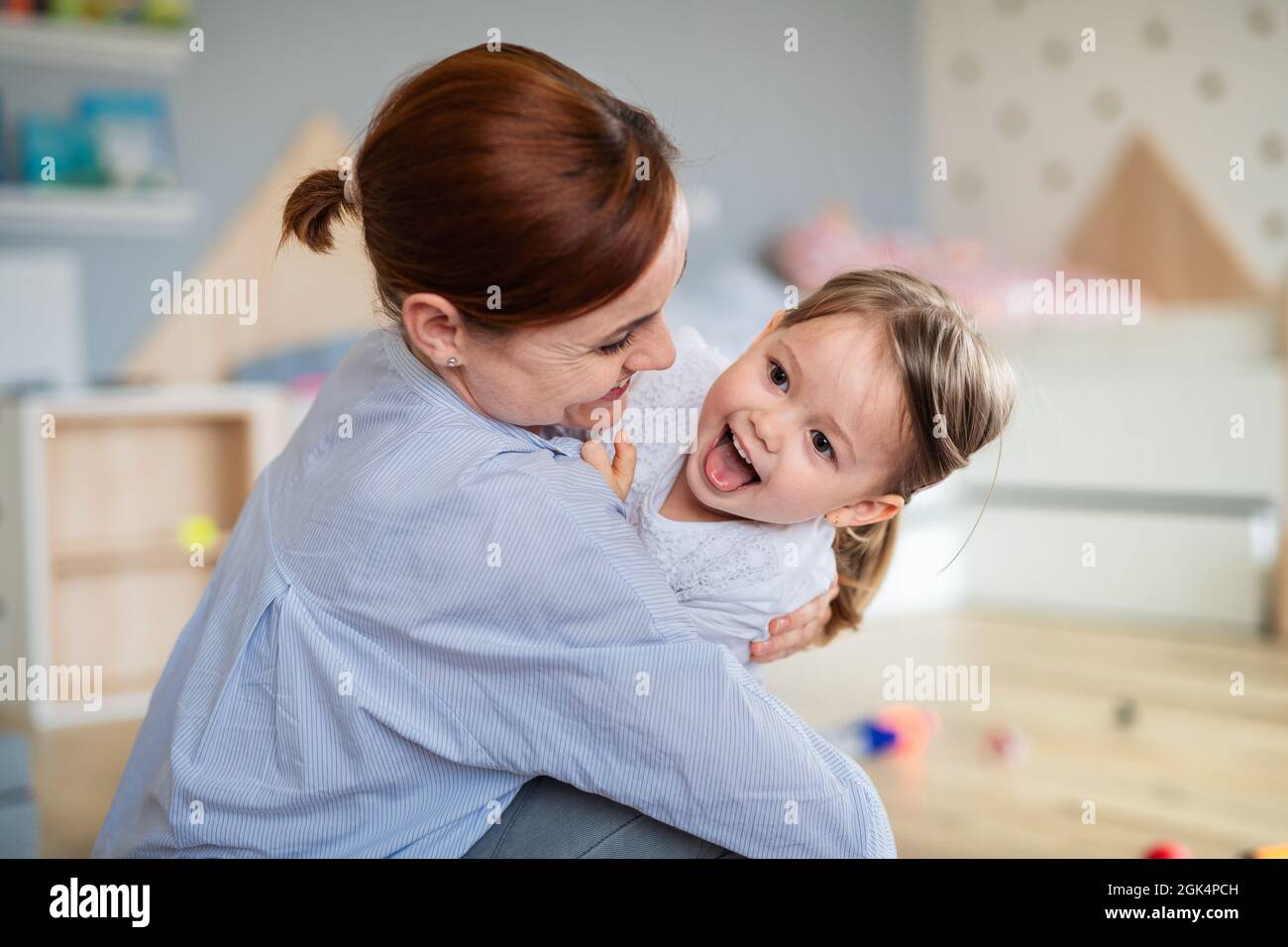 Happy mother with small daughter playing indoors in bedroom, laughing. Stock Photo
