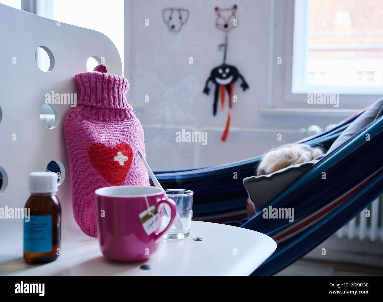 Berlin, Germany. 11th Sep, 2021. A fever thermometer, cough syrup, hot  water bottle and a cup of tea stand on a chair while a child lies in a  hammock in the background.