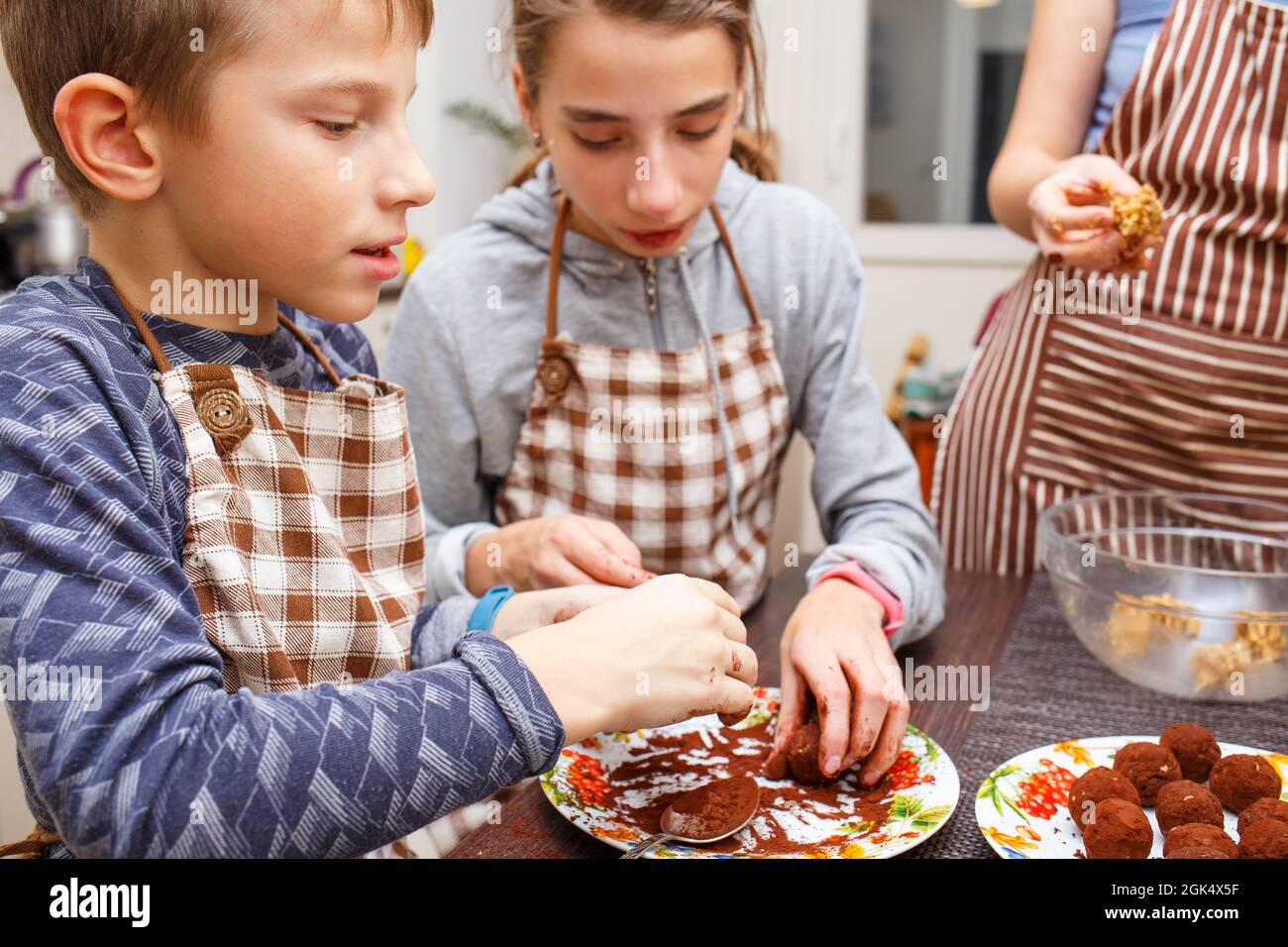 Family cooking sweets at home kitchen. Boy and girl with their mother make healthy sweets with fruits and cocoa. Stock Photo