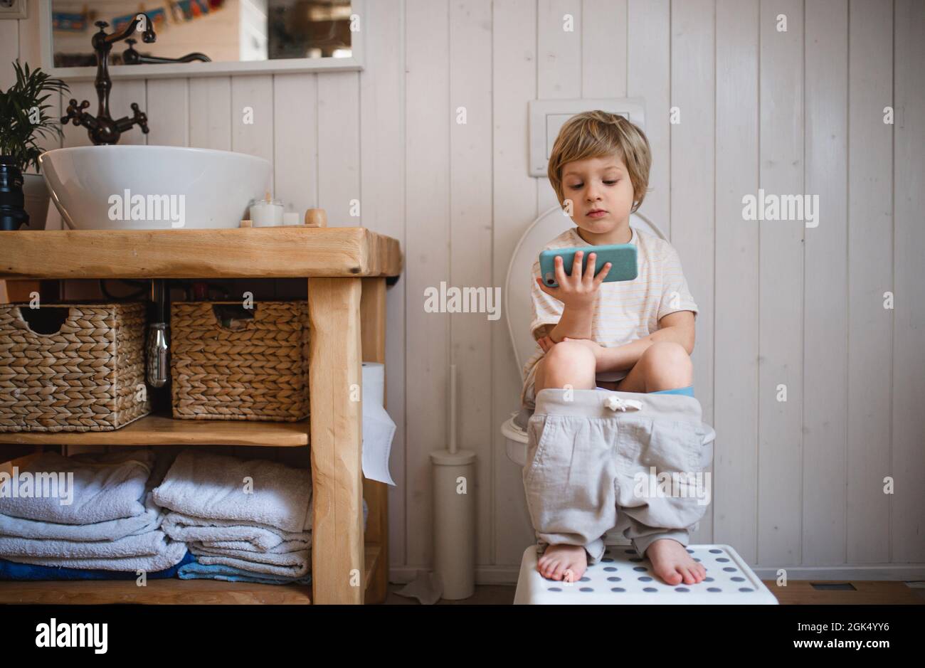 Portrait of cute small boy sitting on toilet indoors at home, using smartphone. Stock Photo