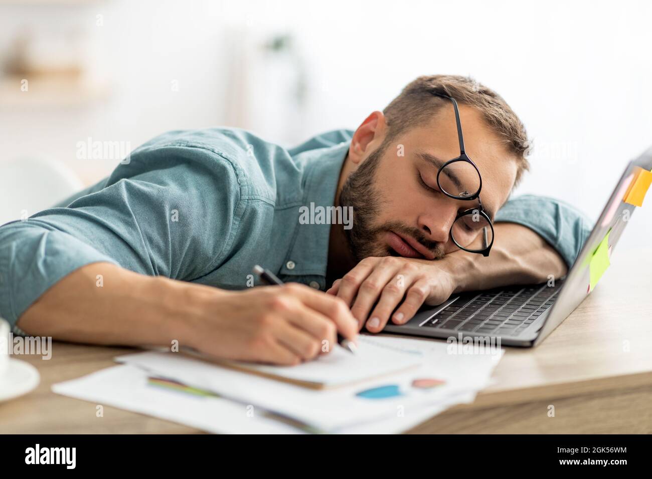 Exhausted millennial man sleeping on his office desk, next to laptop and documents, tired of overworking Stock Photo
