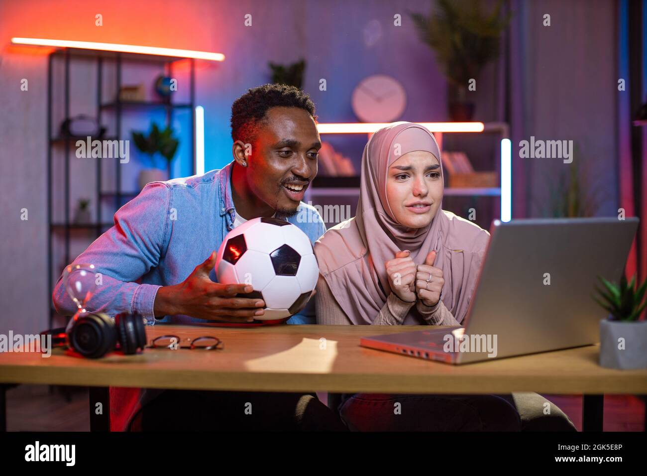 Worried african man and muslim woman watching soccer game on wireless laptop at home. Young multiracial couple enjoying time spending together. Emotions and excitement concept. Stock Photo