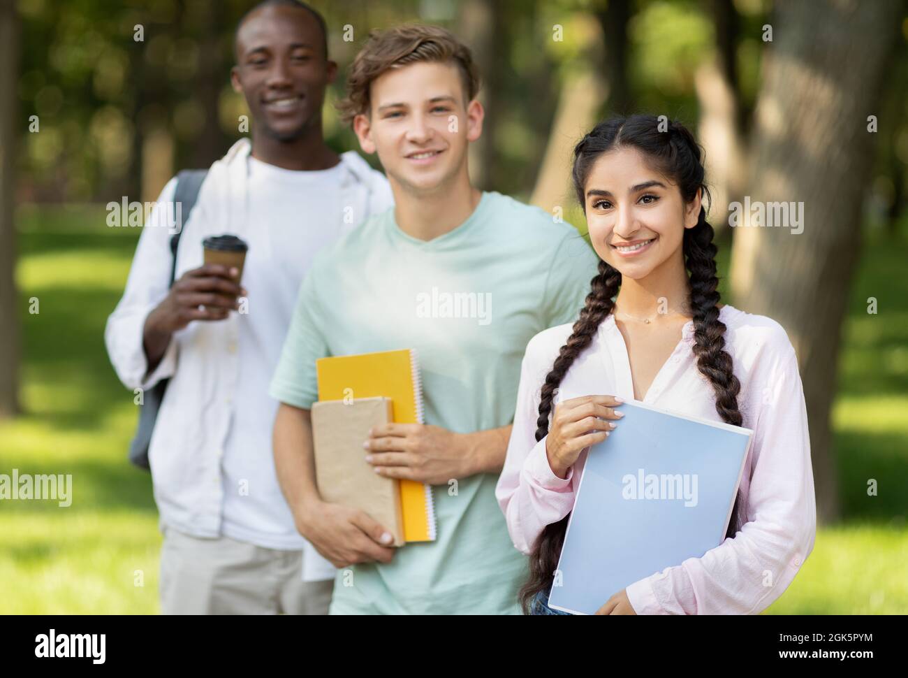University friends. Portrait of multiethnic teens posing outdoors with notepads and books, walking in college campus Stock Photo