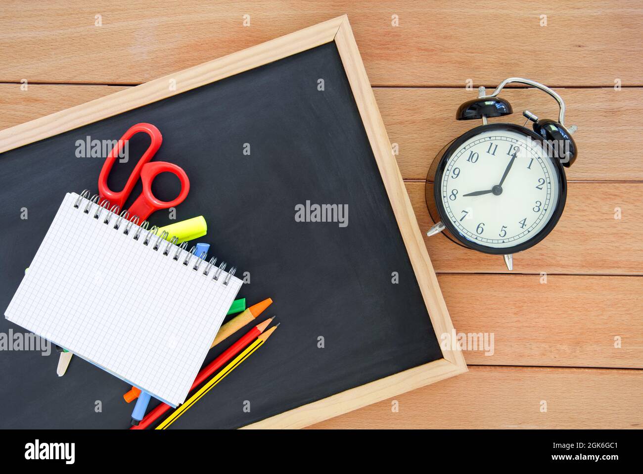 Open notebook and colourful school suplies on a small blackboard lying on a wooden table near an alarm clock. Copy space. Back to school concept. Stock Photo
