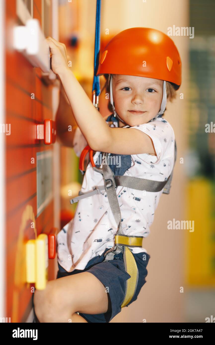 Indoor climbing class for kids. Little boy in red helmet climbing the wall in bouldering center. School boy smiling at the camera and having fun in in Stock Photo