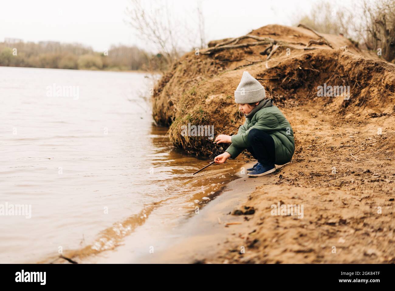 Little child sit on the shore of the lake and playing with water. Happy kid boy playing and having fun outdoors over autumn park background. Stock Photo