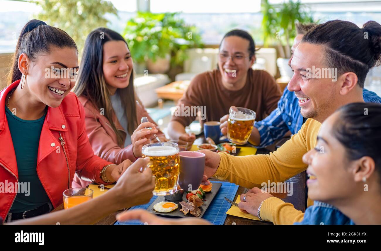 multiethnic group of friends sitting on a table in a bar restaurant making a toast with beers and drinks. diverse people celerating brunch together Stock Photo