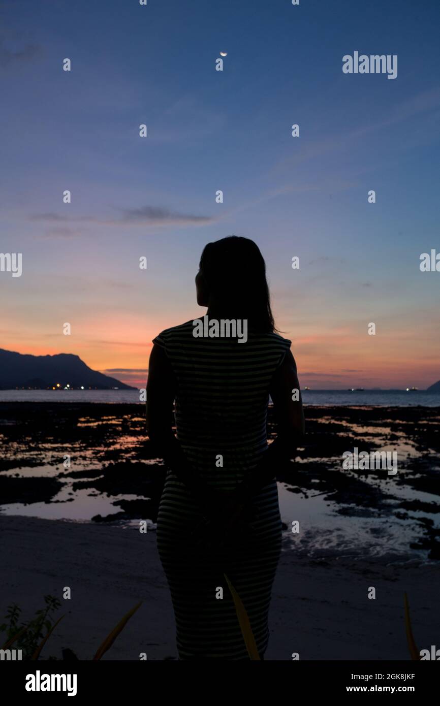 Back view silhouette of unrecognizable female traveler standing on sandy seacoast covered with seaweeds and admiring sunset in Malaysia Stock Photo