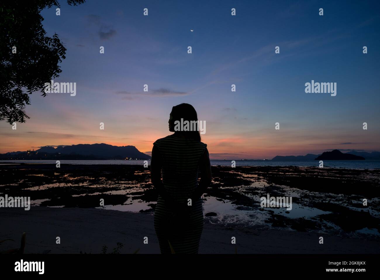 Back view silhouette of unrecognizable female traveler standing on sandy seacoast covered with seaweeds and admiring sunset in Malaysia Stock Photo