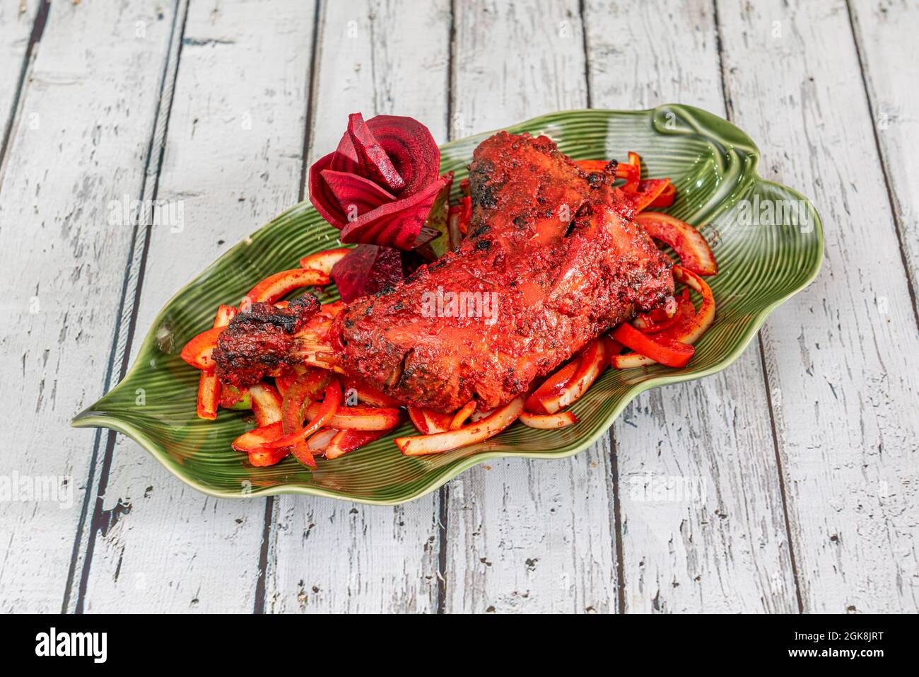 Tandoori chicken thigh seasoned with a lot of typical Indian red sauce on a beautiful green leaf-shaped plate on a white table Stock Photo