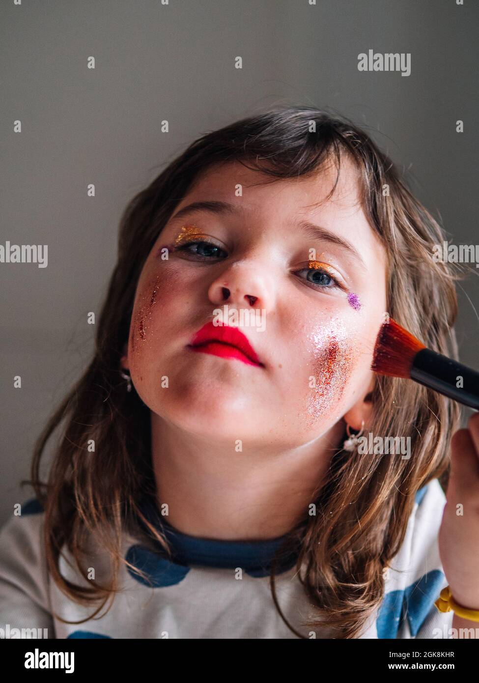 Kid with applicator making up face with assorted cosmetic products in house looking at camera